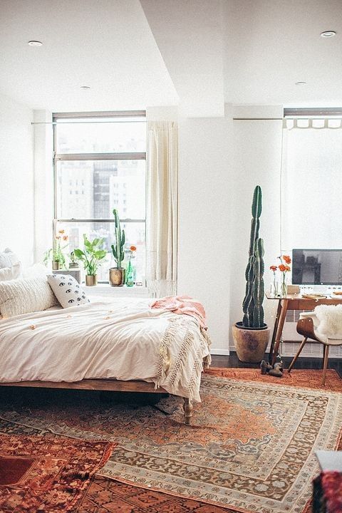 a boho bedroom with layered rugs, a bed with neutral bedding, a desk and a chair, potted plants and decor