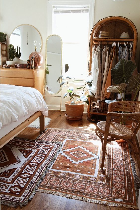 a boho bedroom with layered rugs, a bed with neutral bedding, an arched wardrobe and a chair, a dresser and a mirror and some greenery