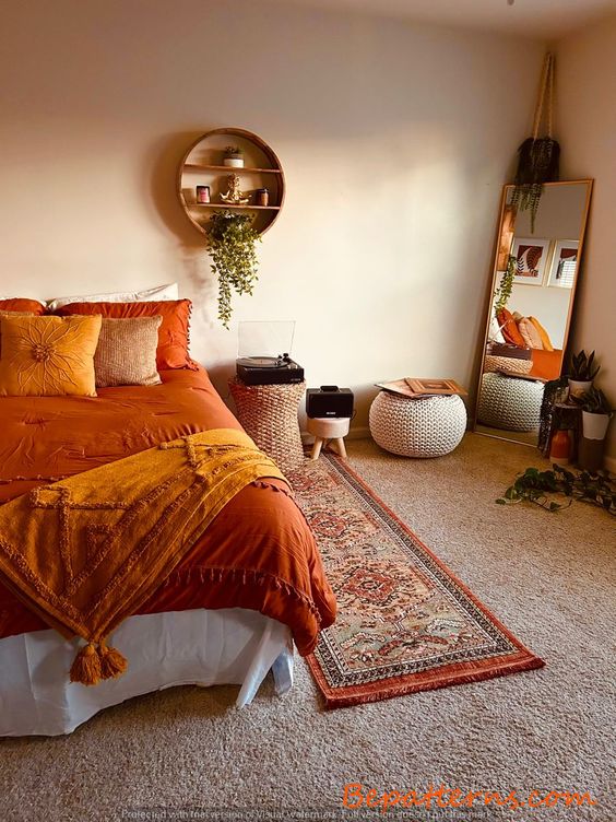 a boho bedroom with layered rugs, a bed with orange bedding, a mirror, poufs and table,s a shelf with decor and some plants