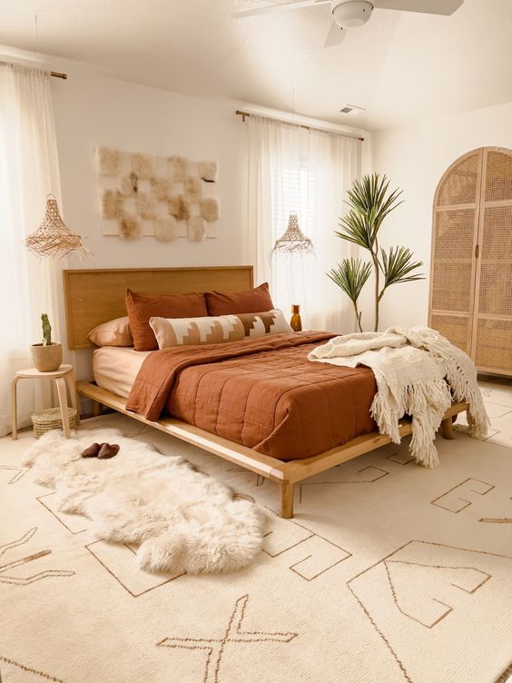 a boho bedroom with layered rugs, a bed with rust bedding, nightstands, a pendant lamp and a potted tree