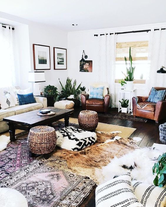 a boho living room with a neutral sofa, brown chairs, layered rugs, various pillows, poufs and various decor
