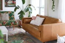 a boho living room with a tan leather couch, a colorful floral loveseat, a bright printed rug, potted greenery and bright artwork