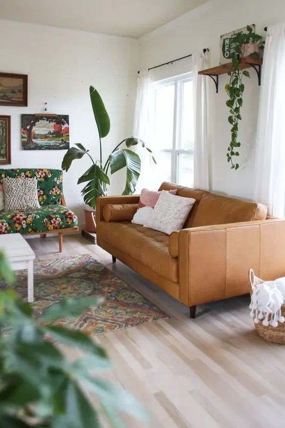 a boho living room with a tan leather couch, a colorful floral loveseat, a bright printed rug, potted greenery and bright artwork