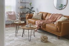 a boho living room with a tan leather sofa, coffee tables, a rattan chair, a shelving unit and a pendant lamp plus a cool big rug