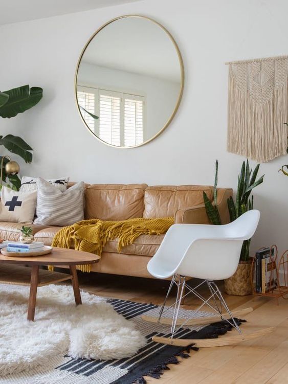 a boho living room with an amber leather sofa and pillows, a chair, macrame, layered rugs and a round mirror