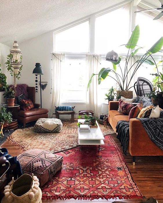 a boho living room with layered rugs, a rust sofa with pillows, a chair, poufs, a coffee table and some potted plants