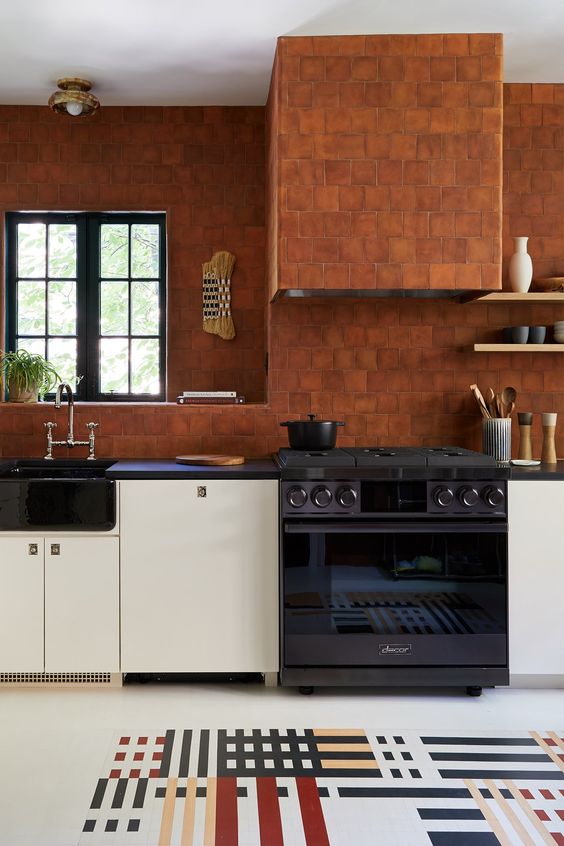 a bold and unique kitchen with white cabinets, black countertops and super bright terracotta tiles on the walls and hood