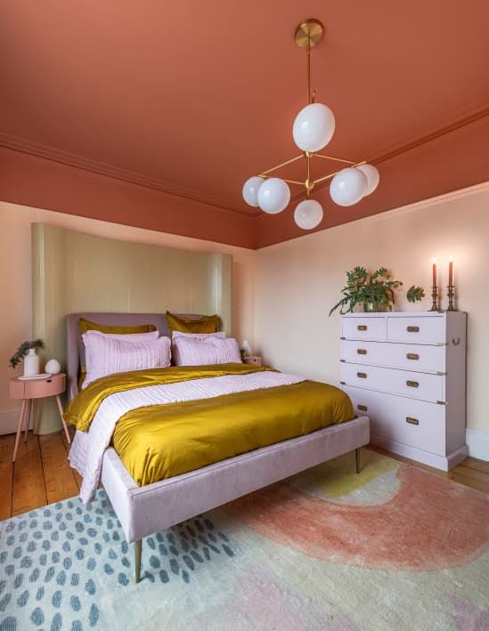 a bold bedroom with a red ceiling, a lilac bed with mustard bedding,a lilac dresser, a bold rug and a pink nightstand