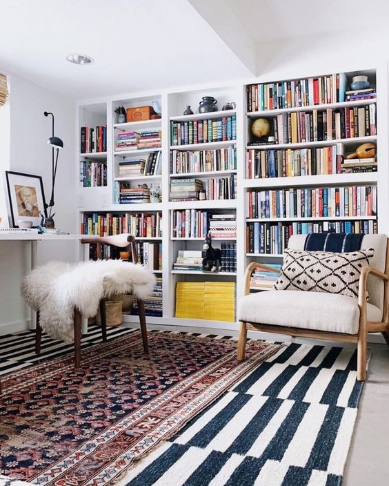 a bold home office with layered rugs, a large bookcase, a desk, some chairs and black lamps