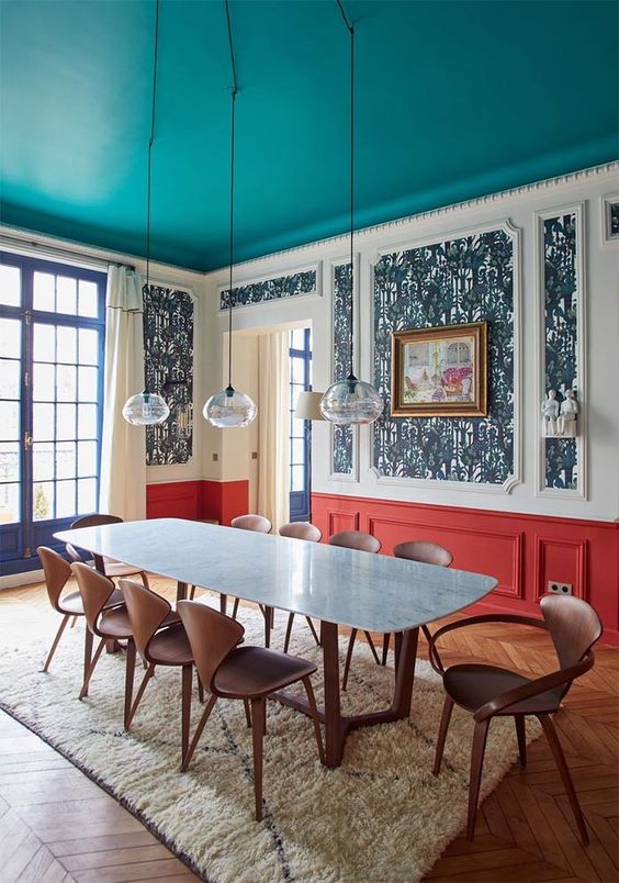 a bright dining room with a turquoise ceiling, bold navy wallpaper and red paneling, a dining table and plywood chairs, pendant lamps