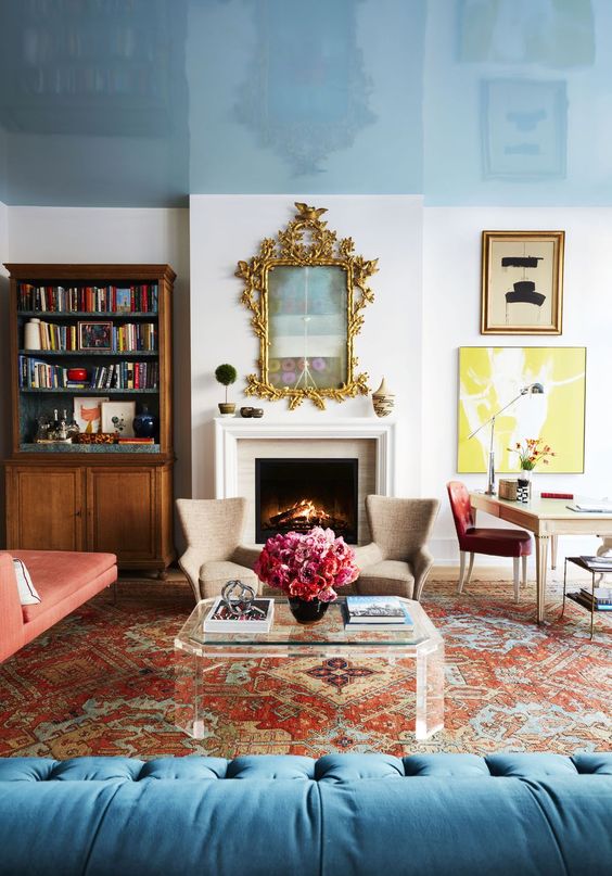 a bright eclectic living room with a glossy blue ceiling, a vintage bookcase, a fireplace, a bold printed rug, a red daybed and a blue sofa