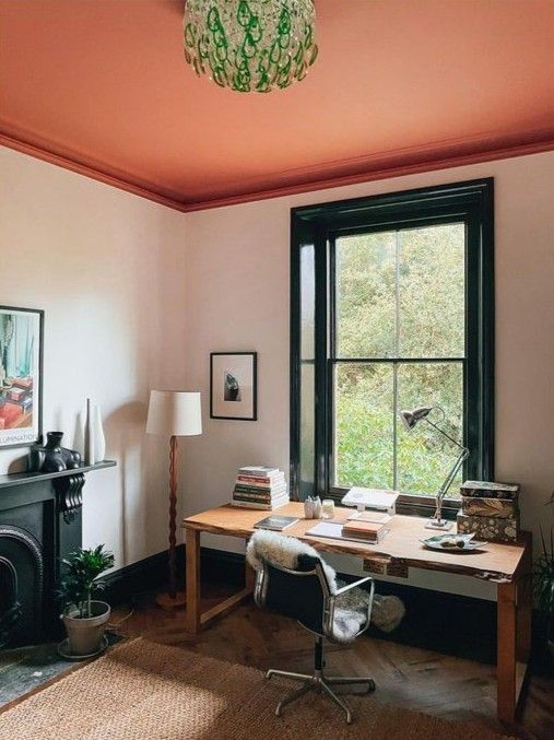 a bright home office with a red ceiling, a black fireplace, a vintage desk, a chair, some lamps and art and a green chandelier