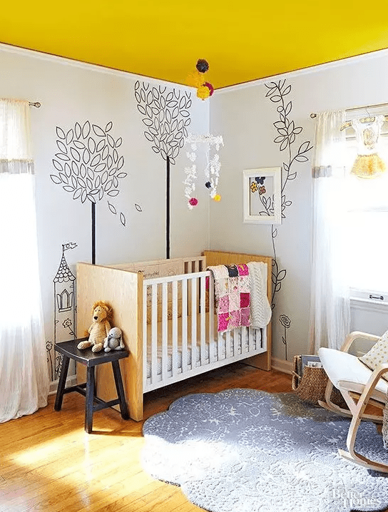 a bright nursery with a bold yellow ceiling, murals on the walls, light stained furniture and a printed rug and some lovely toys