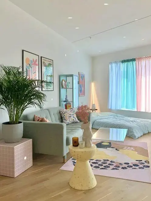 A candy colored living room with a mint sofa and a bed, pastel curtains, a pink side table, a yellow side table, a pastel printed rug and bold artwork