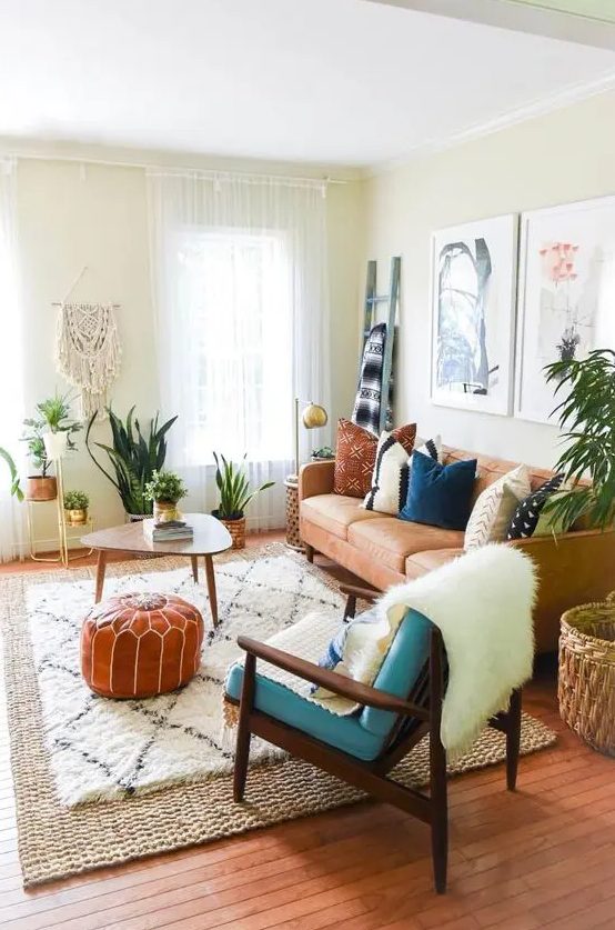 a cozy boho living room with a tan leather couch, a blue chair, a coffee table, potted greenery, a macrame piece and artworks