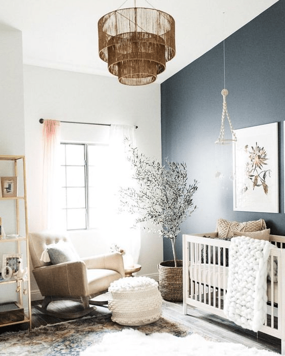 a delicate and cool contemporary nursery with a black accent wall, a white crib, a tan rocker chair, an open shelving unit, a fringe chandelier and a tree