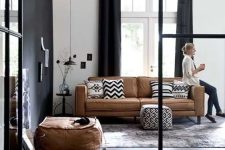 a dramatic living room with a tan leather sofa and a pouf, printed pillows, a black accent wall and black curtains plus a black pendant lamp