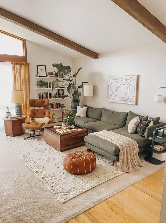 a lovely earthy living room with a green sectional, a stained table, an amber chair and a brown pouf and some bookshelves