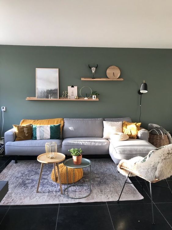 a lovely modern living room with a dark green wall, a grey sofa with pillows, shelves with decor, coffee tables and a chair