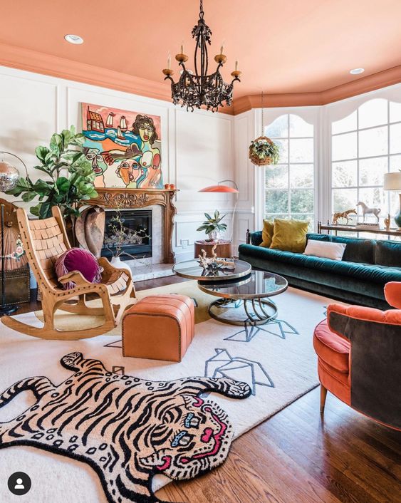 a maximalist living room with a peachy ceiling, a fireplace with a chic mantel, a dark green sofa, a coral chair, a rocker, an orange pouf and fun layered rugs