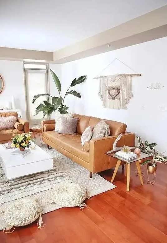 a mid-century modern to boho living room with tan leather furniture, a low white coffee table, woven poufs, a macrame hanging and potted plants
