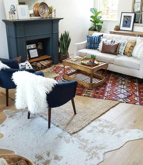 a modern boho living room with layered rugs, a white sofa with pillows, navy chairs, a fireplace, a coffee table and some decor and plants