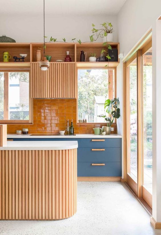 a modern bright kitchen with blue cabinets, a bold yellow backsplash, a fluted hood and a kitchen island, potted greenery and tableware