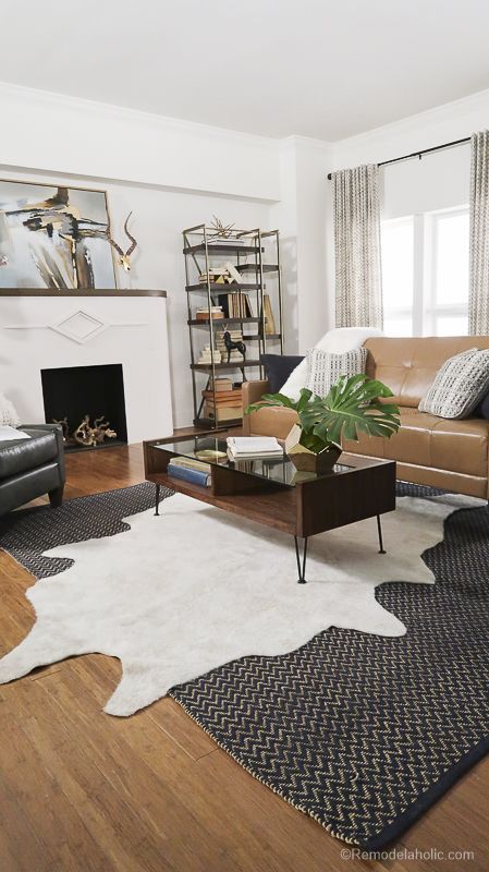 a modern farmhouse living room with a fireplace, a tan leather sofa, layered rugs, a coffee table, a shelving unit and a black chair