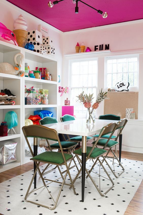 a modern fun dining room with a fuchsia ceiling, a shelving unit, a table, green chairs and lots of quirky and cool decor