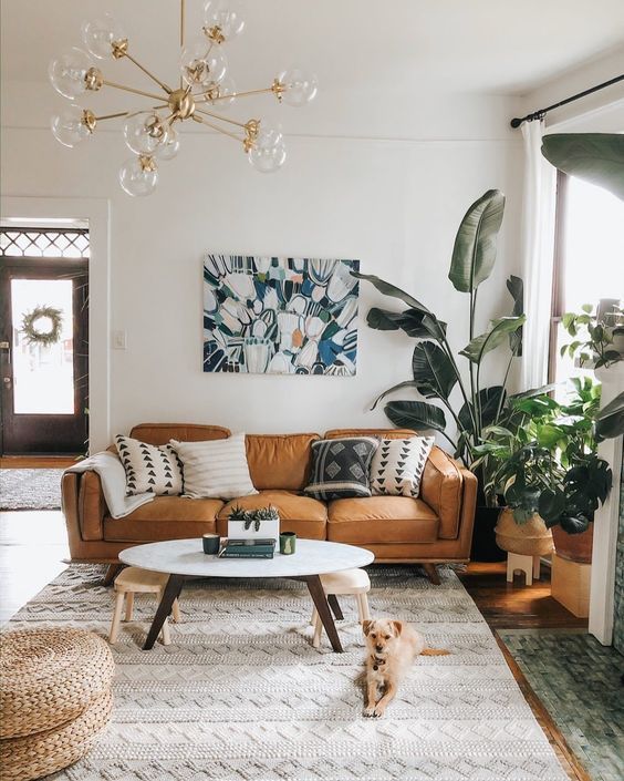 a modern living room with a tan leather ouch, layered rugs, potted plants at the window, an artwork and some pillows