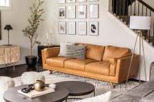 a modern living room with a tan leather sofa, a grey sofa, coffee tables, a credenza, a potted tree and a gallery wall