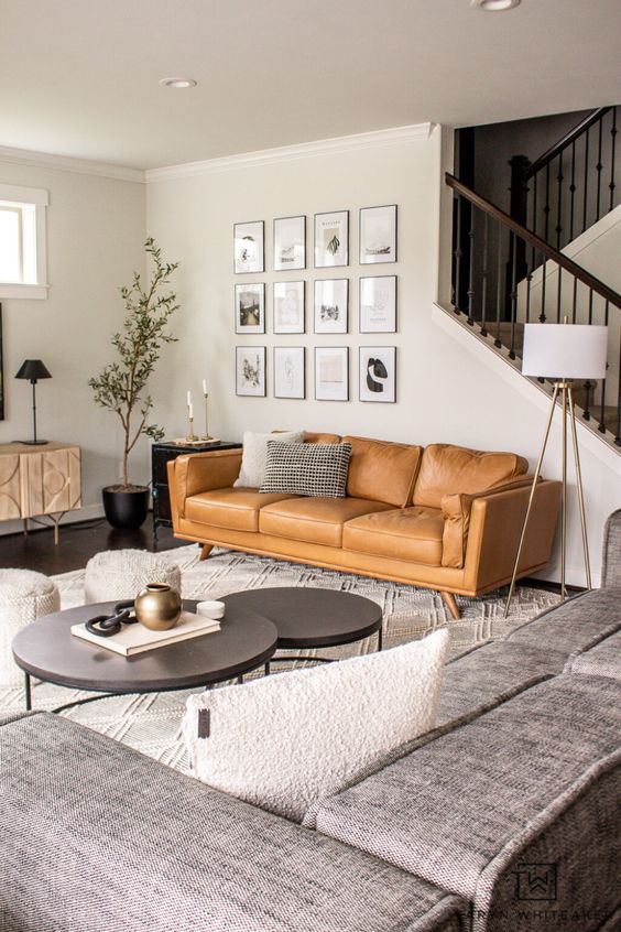 a modern living room with a tan leather sofa, a grey sofa, coffee tables, a credenza, a potted tree and a gallery wall
