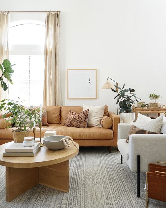 a modern living room with a tan leather sofa, neutral chairs, a stained coffee table, potted greenery, a large rug and a cart