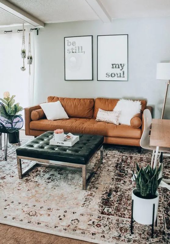 a modern meets boho living room with a tan sofa, a black leather ottoman, a desk and a white chair, a boho rug and potted plants