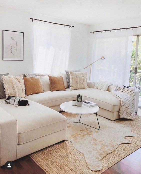a modern neutral living room with a creamy sofa and pillows, layered rugs, a coffee table and an artwork
