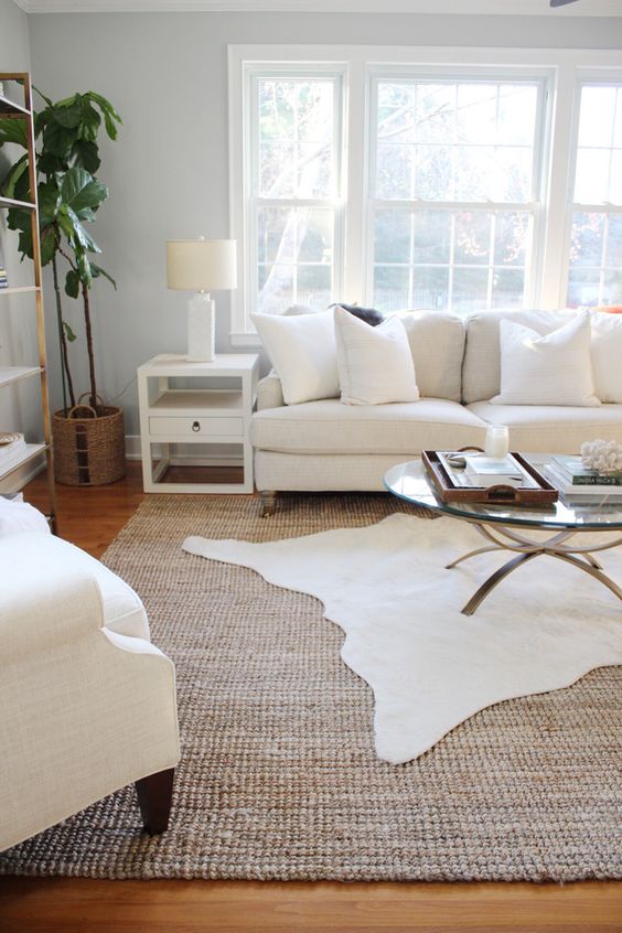 a modern neutral living room with layered rugs and white seating furniture, a glass coffee table and a potted tree