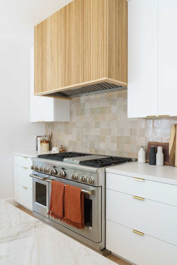a modern white kitchen with a tan and grey tile backsplash, white countertops and a fluted hood