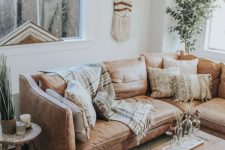 a neutral boho living room with a tan leather sofa, a chest, a side table, potted greenery, printed pillows and macrame