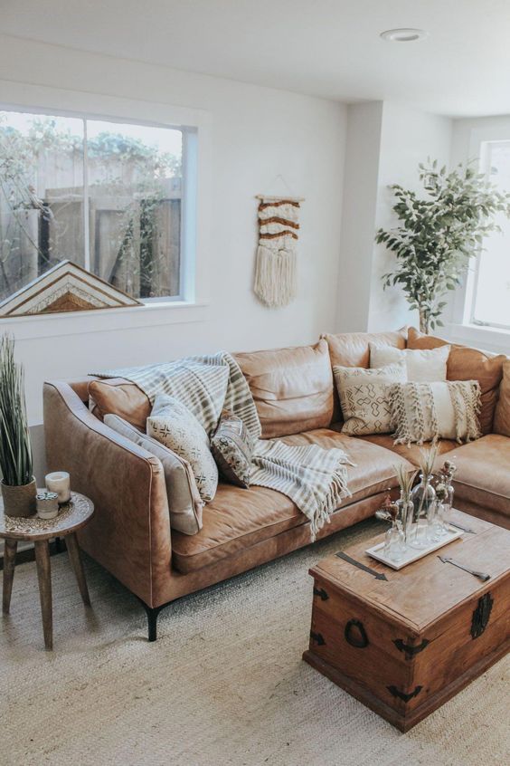 a neutral boho living room with a tan leather sofa, a chest, a side table, potted greenery, printed pillows and macrame