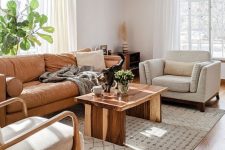 a neutral modern living room with a tan leather couch, white chairs, a coffee table, a printed rug and some greenery