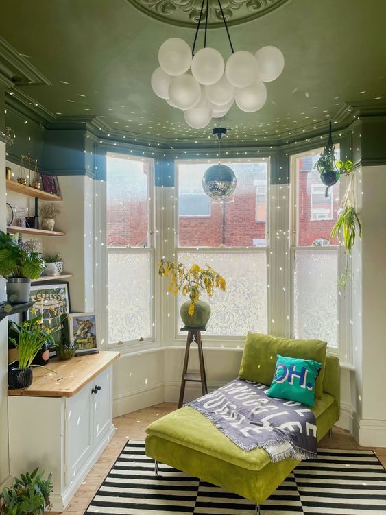 a pretty bay window space with a green ceiling, a cabinet and shelves with art, a green daybed and a striped rug plus a disco ball
