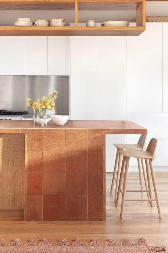 a sleek white kitchen with a kitchen island clad with terracotta tiles, open shelves, tall stools and some blooms