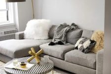 a small and chic living room with dove grey walls, a grey sectional, a striped coffee table, a faux fur stool, a shelf with a potted plants and an artwork