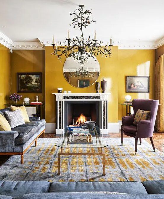 a sophisticated living room with mustard walls, a grey sofa and a mauve chair, a fireplace, a round mirror, a chic chandelier