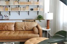 a stylish living room with bookshelves that take a whole wall, an amber leather sofa and an ottoman, an acrylic table and greenery