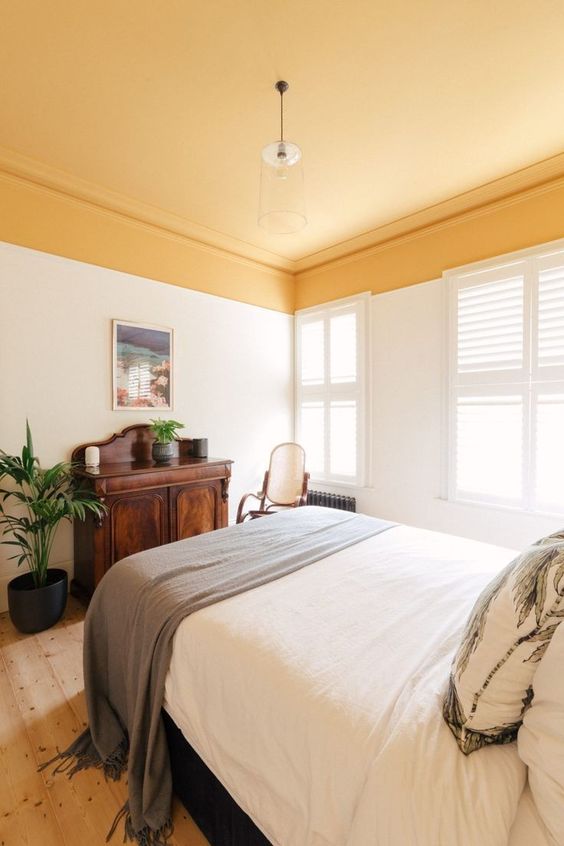 A warm and welcoming bedorom with a yellow ceiling, a bed with neutral bedding, a dark stained cabinet, a chair and potted greenery