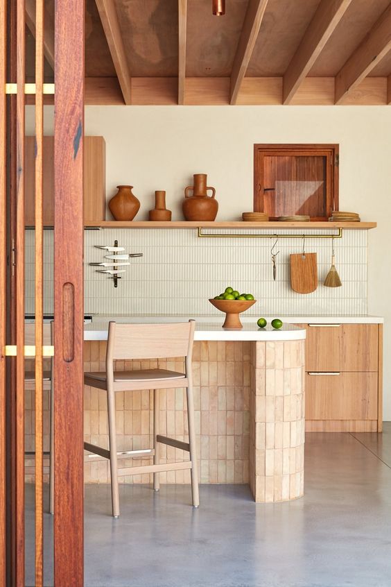 a welcoming earthy kitchen with a wooden ceiling, stained cabinets, a kitchen island clad with terracotta tile, stools and shelves