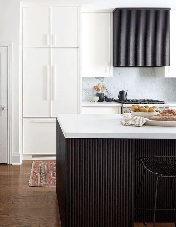a white kitchen with a white backsplash and countertops,a  black fluted kitchen island and a hood is chic and cool