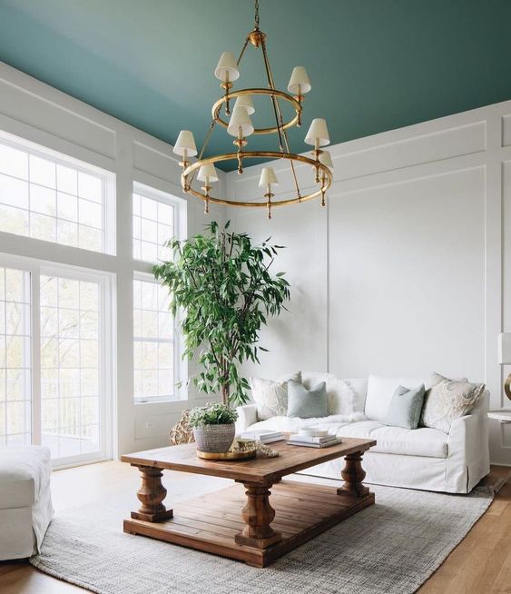 an airy living room with a green ceiling, a white sofa, a stained vintage table, a tiered chandelier and a rug