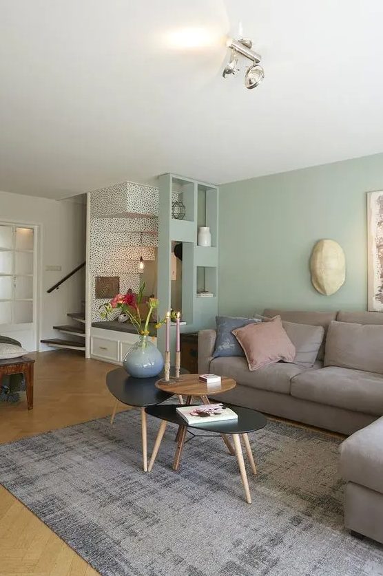 an airy living room with light green walls, a grey sectional, some coffee tables, a grey rug and cool decor